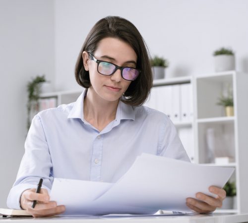 Young serious female accountant in eyeglasses and formalwear reading papers